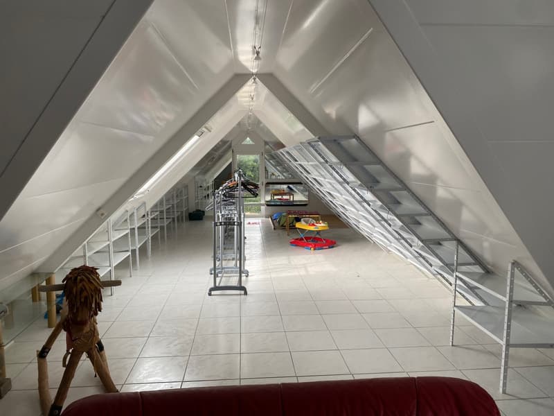 The image shows a spacious attic space with a slanted ceiling and white walls. The floor is tiled, and the room is filled with various storage items, including shelves, a ladder, and some toys. The space appears to be used for storage and organization.