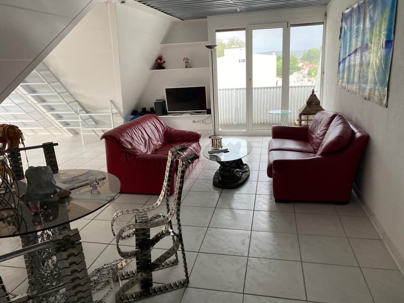 Spacious living room with large windows, red leather sofa and armchair, glass coffee table, TV stand, and decorative items on shelves. The room has a tiled floor and opens up to a balcony or terrace.