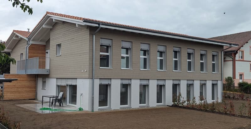 Modern two-story building with grey siding, large windows, and a balcony on the upper level. The ground floor features a patio area with a table and chairs.