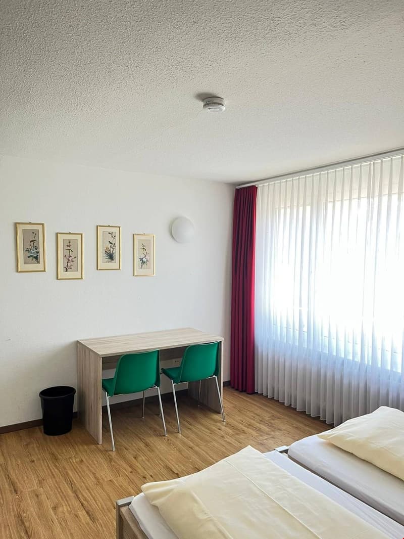 Bedroom with hardwood flooring, white walls, framed artwork, a wooden desk with green chairs, and a large window with sheer curtains.