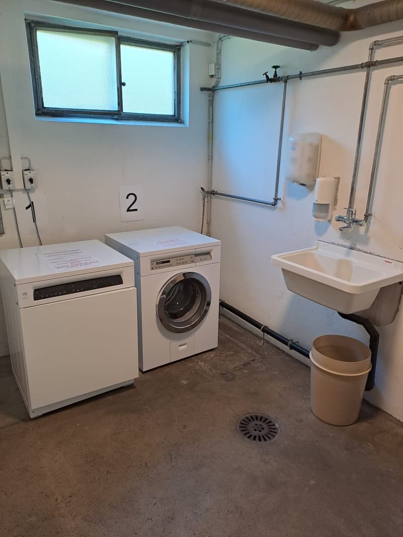 The image shows a laundry room with a washing machine, a dishwasher, a sink, and a window. The room has concrete flooring and exposed pipes on the walls.