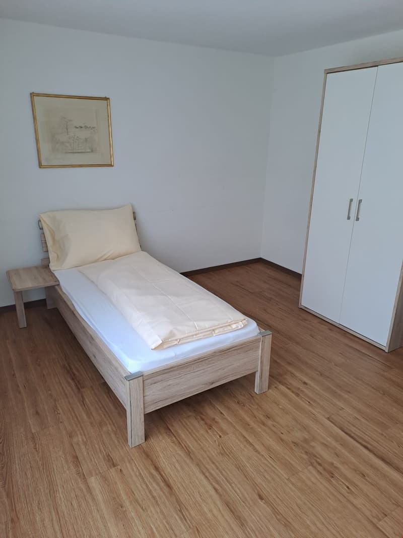 The image shows a simple bedroom with a single bed, a wooden nightstand, and a white wardrobe. The room has light-colored walls and a hardwood floor.