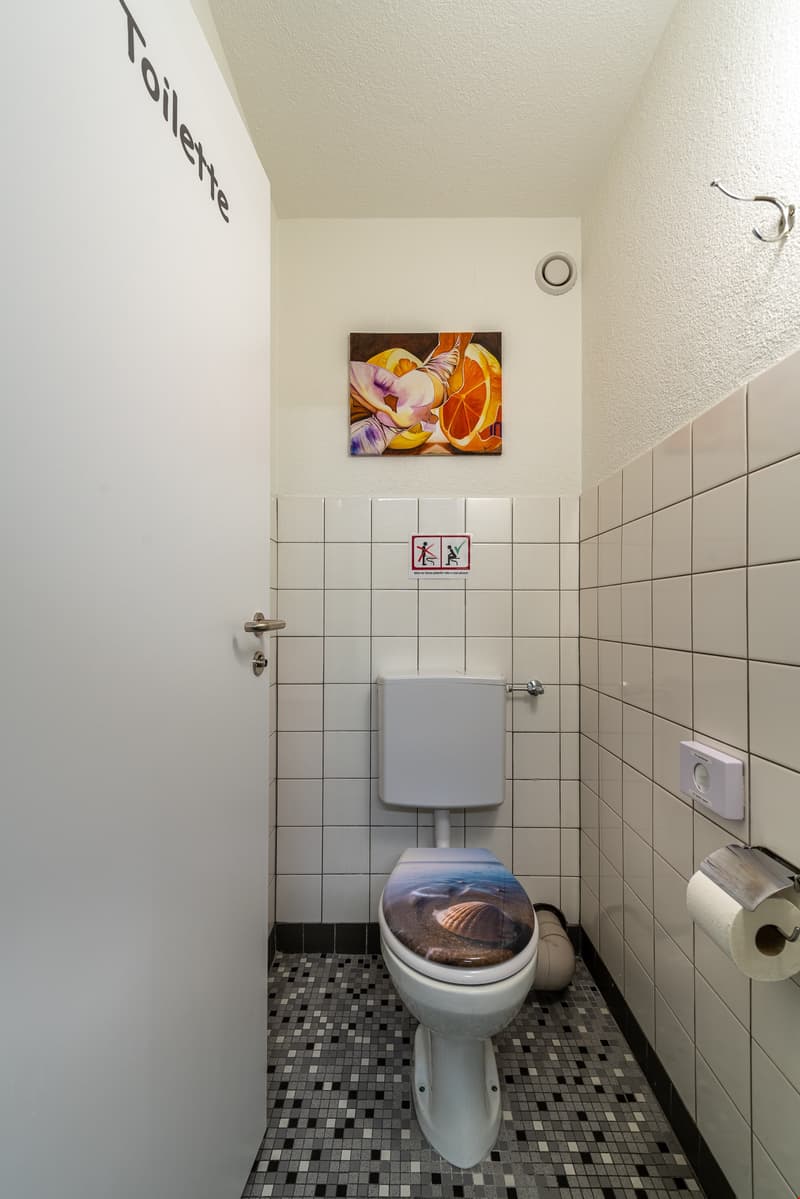 Tiled bathroom with white walls, black and white mosaic floor tiles, toilet, and a framed artwork on the wall.