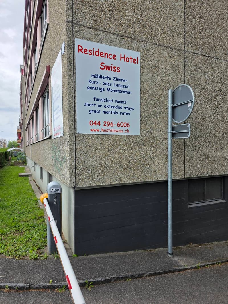 Exterior view of a building with a sign reading "Residence Hotel Swiss" advertising furnished rooms for short or extended stays with great monthly rates. The building has a textured concrete facade and a small garden area visible to the left.
