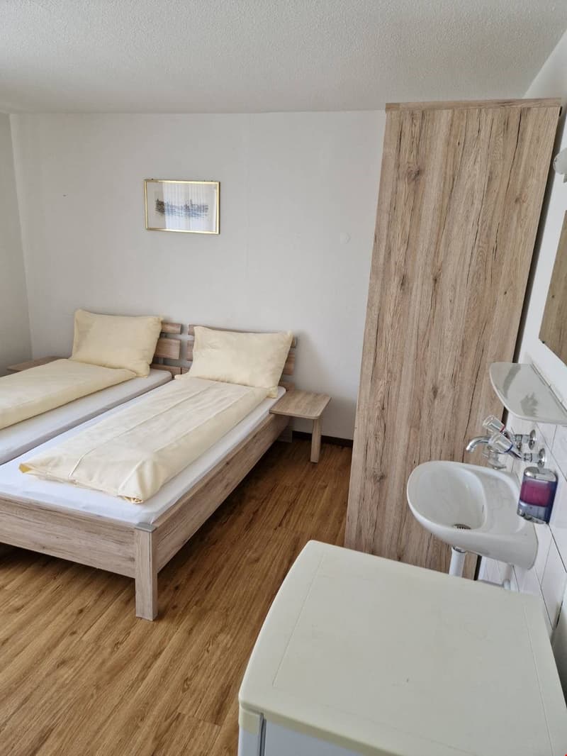 Spacious bedroom with wooden furniture, including a bed frame and side table. The room has white walls and a hardwood floor. There is a small framed artwork on the wall.