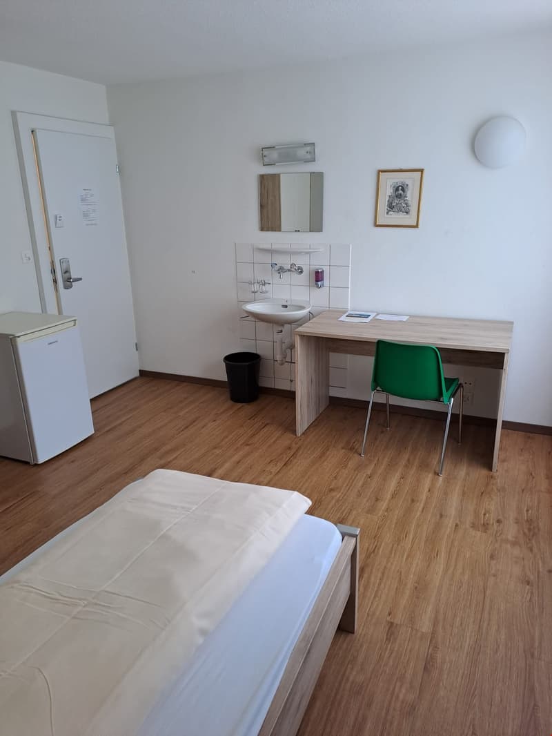 The image shows a studio apartment with a bed, a desk with a chair, a sink and vanity area, and a small refrigerator. The room has a hardwood floor and white walls. There is a framed artwork on the wall and a mirror above the sink. The apartment appears t