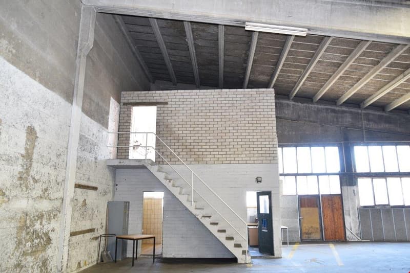 The image shows a large, open industrial space with a high ceiling, exposed wooden beams, and brick walls. There is a staircase leading up to a second level, and a door leading to another room or area. The space appears to be in a state of disrepair or re