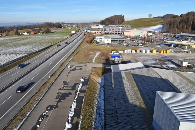 The image shows an aerial view of a highway surrounded by commercial and industrial buildings, with a snowy landscape in the background. The highway has multiple lanes and is busy with traffic. There are several large buildings and warehouses visible, as