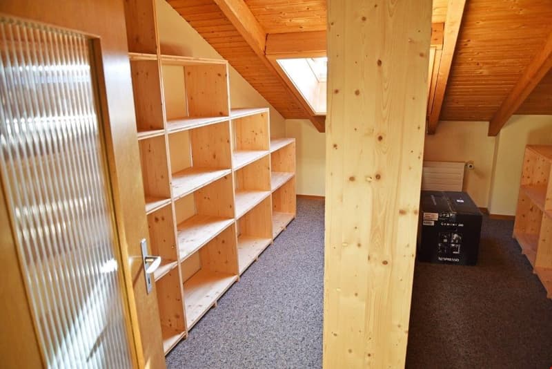 Attic space with wooden shelving units, wooden beams, and carpeted floor. The room appears to be used for storage or additional living space.