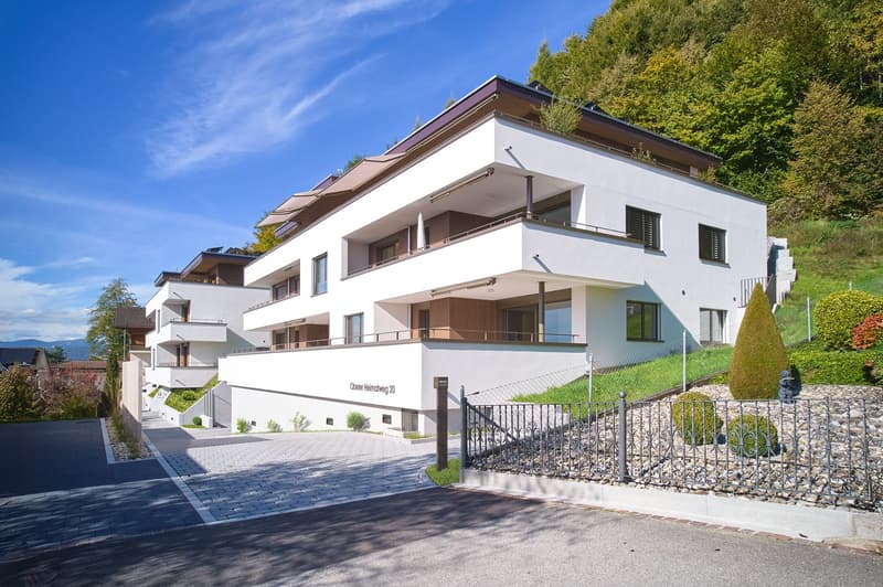 Modern, white building with several balconies and windows, located on a hill, surrounded by greenery.