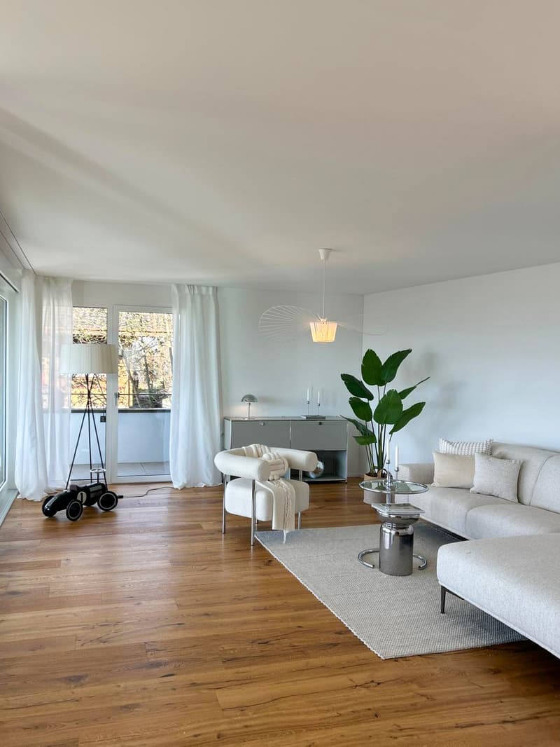 open living room with wooden floors, white walls, glass windows with curtains, sliding door, large couch, ottoman, side table, lamps, coffee table, potted plant, toy car