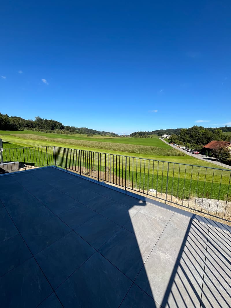 The image shows a large outdoor terrace or balcony overlooking a scenic rural landscape. The terrace has a blue tiled floor and a metal railing. Beyond the railing, there is a vast green field with trees and hills in the distance under a clear blue sky.