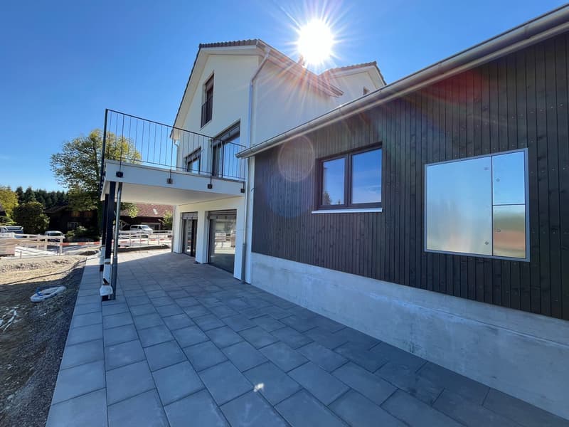 2-story house with white exterior walls, black wooden siding, large windows, and a balcony on the second floor. The house has a paved patio area in the front with a tiled floor.