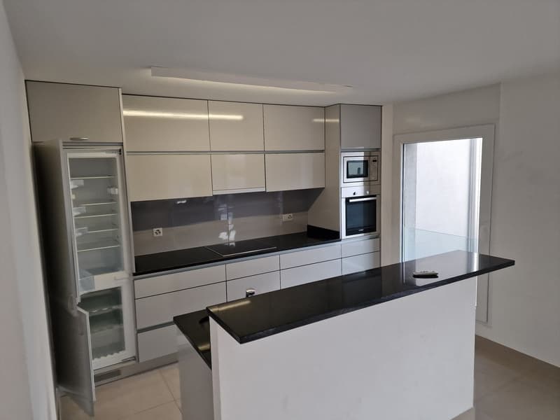Modern, open-concept kitchen with white cabinets, black countertops, built-in appliances including a dishwasher, and a breakfast bar or island
