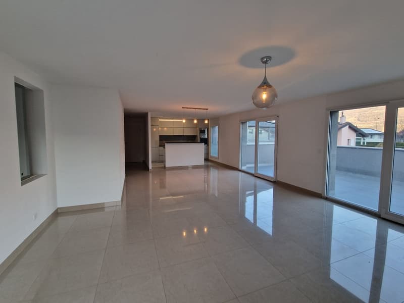 Spacious open-plan living area with large windows, modern kitchen, and sliding glass doors leading to a balcony or terrace. The room has a sleek, minimalist design with high-quality tile flooring and recessed lighting.