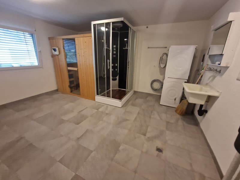 The image shows a studio apartment with a tiled floor, a shower enclosure, a washing machine, and a refrigerator. There is also a wooden cabinet and a desk visible.