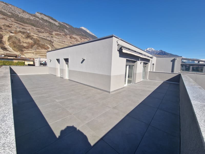 A modern, two-story building with a flat roof and large windows. The exterior is painted in a light gray color. The building has a spacious terrace or balcony area with a tiled floor. The surrounding landscape includes mountains in the background.
