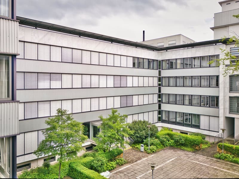 Large, modern building with multiple floors, white exterior, many windows, courtyard, parking area.