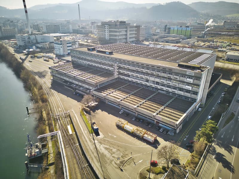 Large multi-story industrial building with a flat roof, surrounded by other commercial and industrial buildings. The building has a rectangular shape and appears to be used for manufacturing or warehousing purposes. There are several vehicles parked in th
