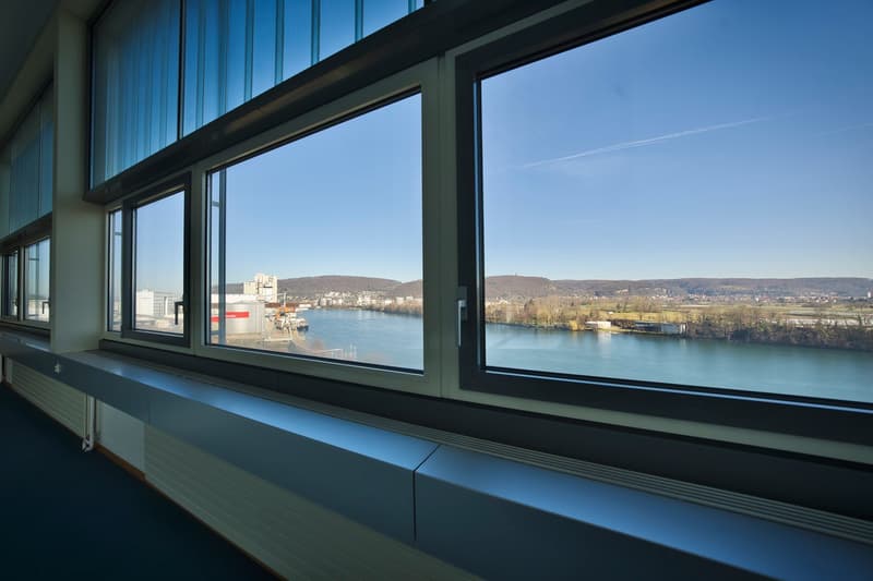 Large windows with a scenic view of a river, cityscape, and mountains in the background. The interior appears to be a modern office or commercial space with a sleek, minimalist design.