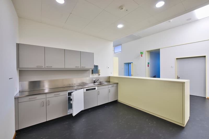 Modern kitchen with stainless steel appliances, including a dishwasher, and gray cabinets. The kitchen has a counter and sink area with a window. The room also has a tiled floor and recessed lighting in the ceiling.