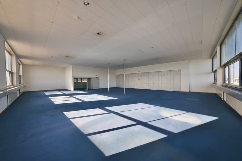 Large open space with blue carpeted floor, white walls, and ceiling lights. The room appears to be a gymnasium or exercise facility with lockers visible along the walls.