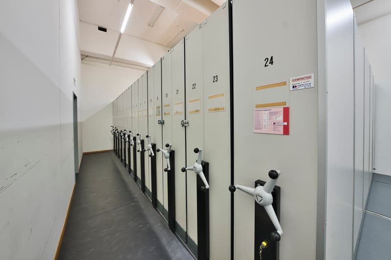The image shows a hallway with a row of numbered storage lockers on both sides. The lockers have locks and handles, and there is a sign indicating the locker numbers. The floor is a dark tile or concrete material.