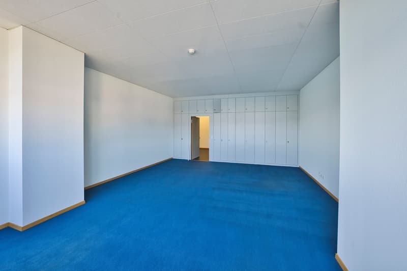 Large open room with blue carpeted floor, white walls, and built-in lockers or storage units along one wall. The room appears to be a communal or shared space, possibly in an office, school, or other institutional setting.