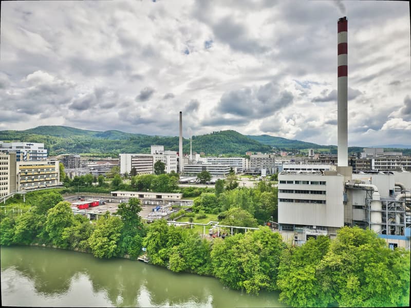 The image shows a cityscape with a mix of industrial and commercial buildings, including a tall chimney stack with red and white stripes. The city is surrounded by green hills and a river runs through the center. There appears to be a parking area or lot