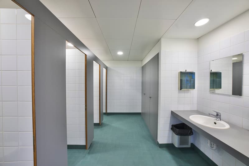 The image shows a bathroom hallway with white tiled walls, a sink with a mirror, and a green tiled floor. The hallway has multiple doors leading to other rooms.