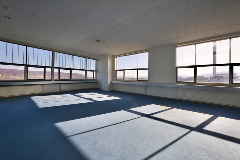 Large open office space with floor-to-ceiling windows offering a panoramic view of the city skyline. The room has a blue carpeted floor and a white ceiling with recessed lighting. The space appears to be empty and ready for use.