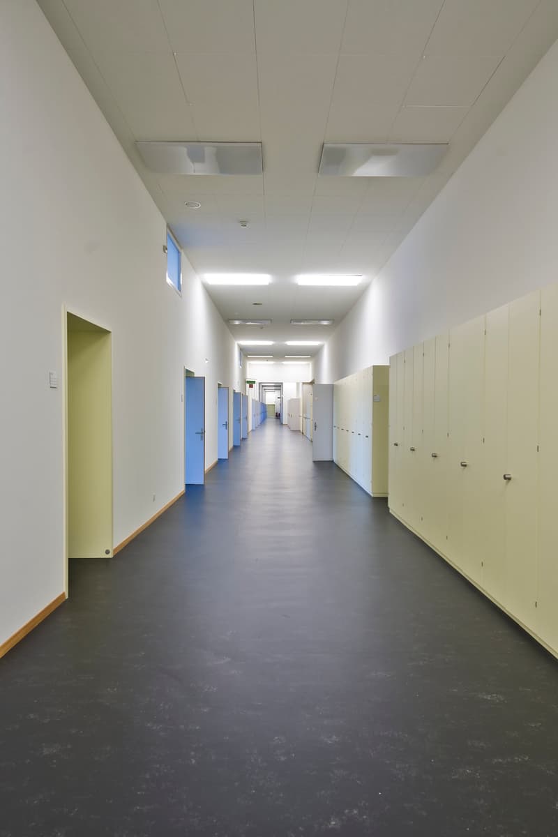 Long, well-lit hallway with white walls, gray flooring, and lockers or storage units lining the walls on both sides.