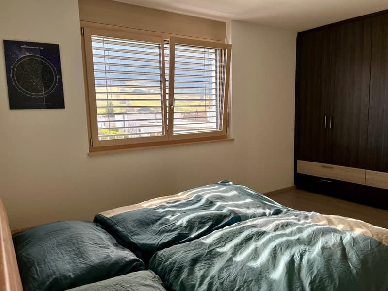 The image shows a bedroom with a large window, a dark wooden wardrobe, and a bed with a crumpled dark bedspread. There is a framed star chart on the wall.