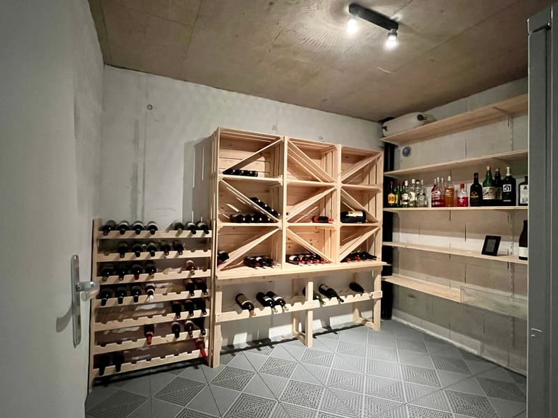 Wine cellar with wooden shelves and gray tiled floor, bottles of wine and other beverages, lights on the ceiling.