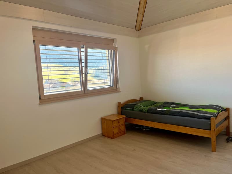 The image shows a bedroom with a wooden bed frame, a bedside table, and large windows overlooking a scenic outdoor view. The room has a neutral color scheme with light walls and floors.