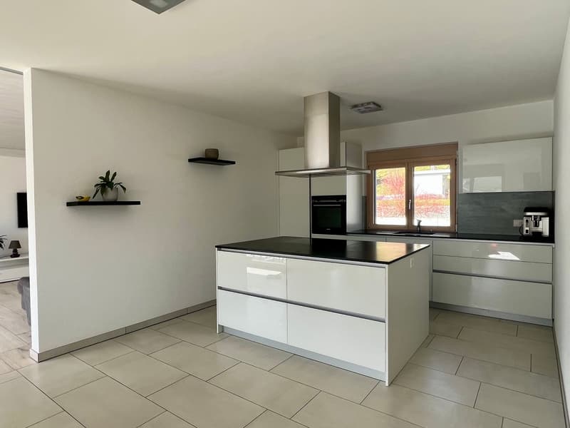 Modern, open-concept kitchen with white cabinets, black countertops, and stainless steel appliances including a dishwasher. The kitchen has a central island with drawers and a built-in oven and cooktop. The room has large windows providing natural light.