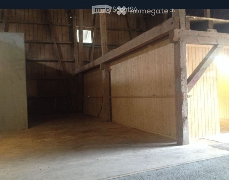 The image shows a spacious attic or storage area with wooden beams and walls. The space appears to be unfinished, with a concrete floor and some storage items visible. There is a small window providing natural light to the space.