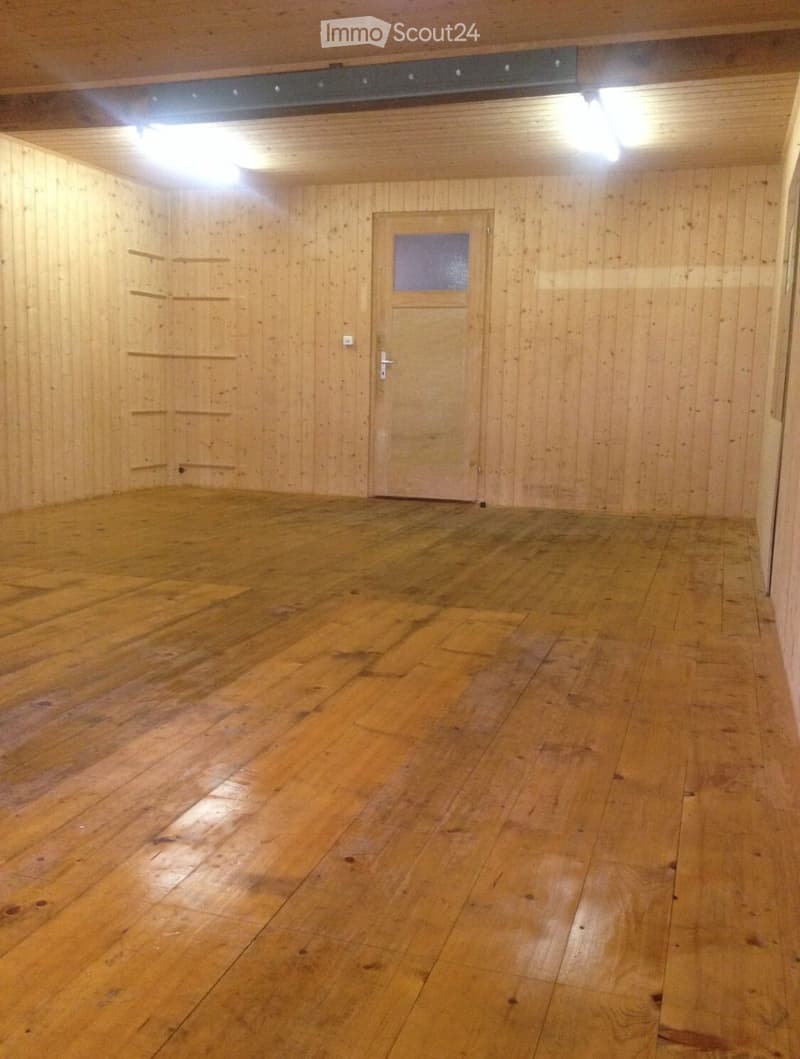 This image shows a large, open room with wooden walls and floors. The room has a single door and is illuminated by overhead lighting. The wooden floor appears to be in good condition, with some visible wear and tear. The walls are made of wood paneling, c
