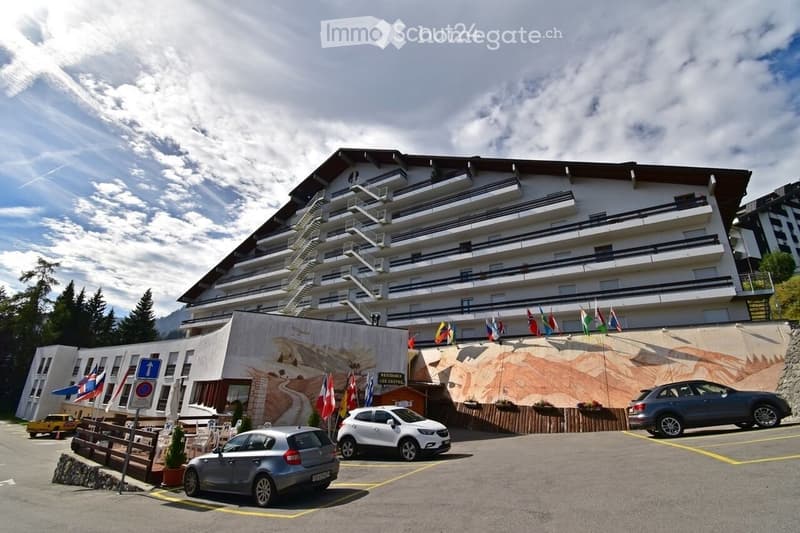 multi-story building, several cars parked in front, flagpoles, mountain-themed mural on the wall