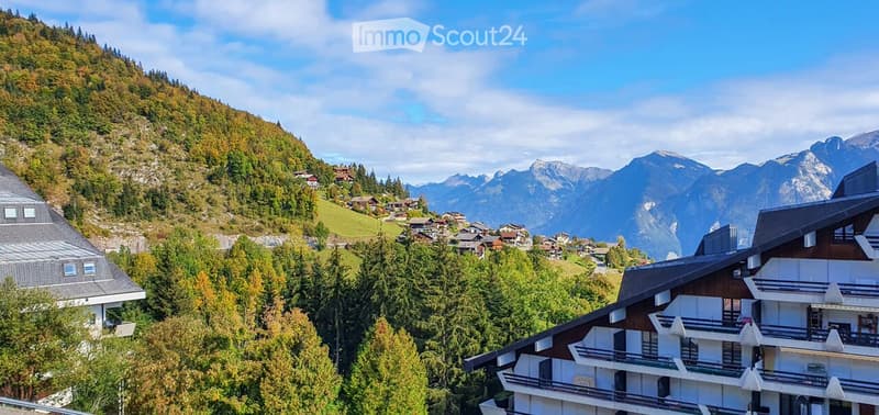 A view of several houses nestled on a hillside in a mountainous region, surrounded by trees and featuring a distant view of the Alps