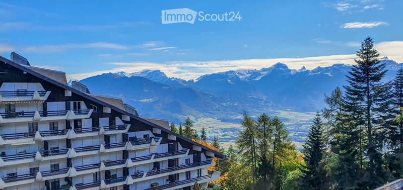 Modern building with multiple balconies, surrounded by trees and mountains
