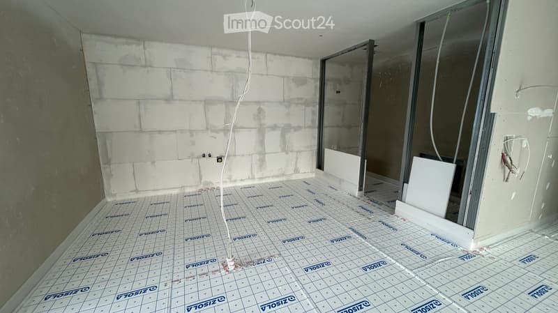 Empty room with unfinished walls and floor, showing exposed wiring and insulation. The floor has a protective grid pattern, and the walls are partially covered with white tiles.