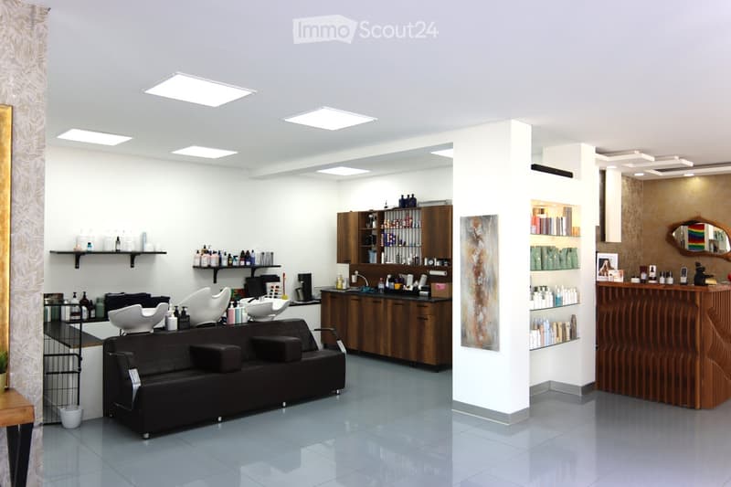 Modern interior, white walls, tile flooring, black leather couch, wooden cabinet, shelves with bottles, and various hair styling equipment.