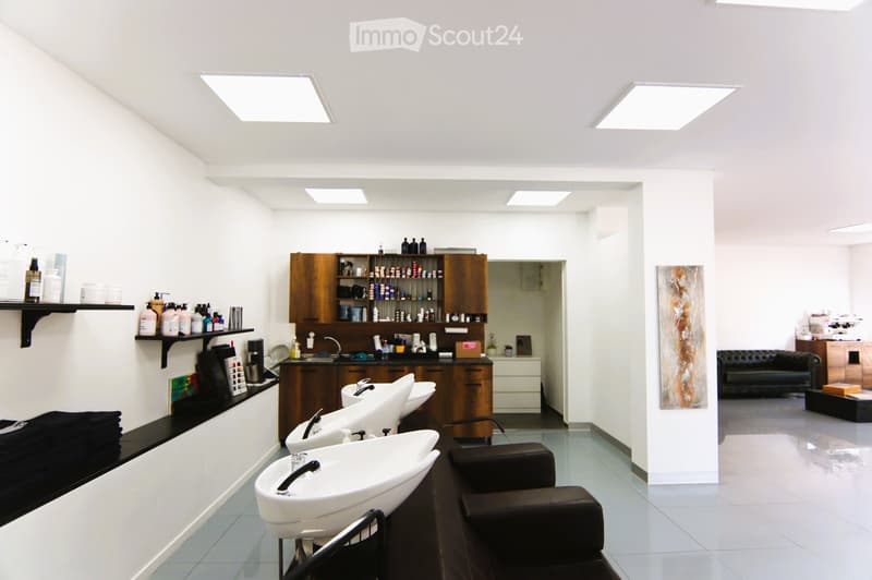 Interior of a salon with white walls, three washing sinks, a wooden cabinet, leather chairs, a countertop, a black sofa, white tiles, square lights, shelves with hair products, and a picture frame on the wall.
