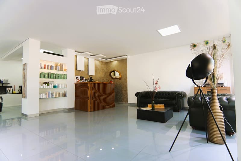 Open floor plan, white tiled flooring, white walls, wooden desk, black couch, vase with feathers, standing lamp, wooden coffee table, shelves with products