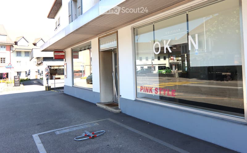 Exterior view of a building with glass windows and a sign reading 'OKN PINK STYLE', situated along a road with cars parked and a storefront entrance. Street includes a parked bicycle stand.