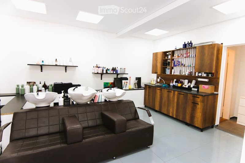 Commercial space with a counter, sink, cabinets, shelves, salon chair, three basins, bottles, and bottles on shelves, lamps, and tile flooring.