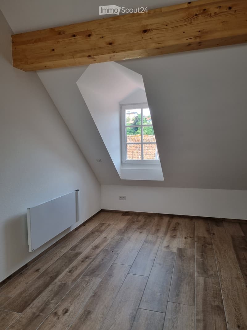 empty attic room, wooden floor, white walls, wooden beam, slanted ceiling, window, radiator