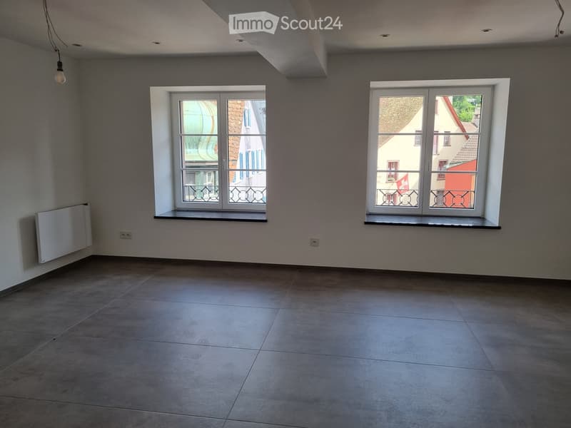 Empty room with white walls, gray tiles, two windows, power outlets, window sills, ceiling light fixture.