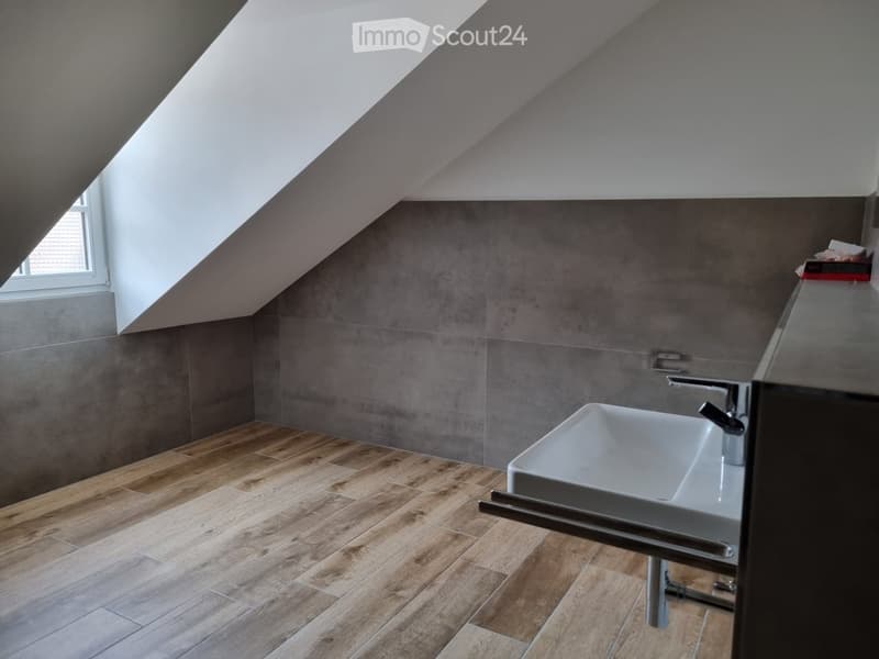 white sink, grey tiles on wall, wooden tiles on floor, white walls, metal faucet, window on ceiling